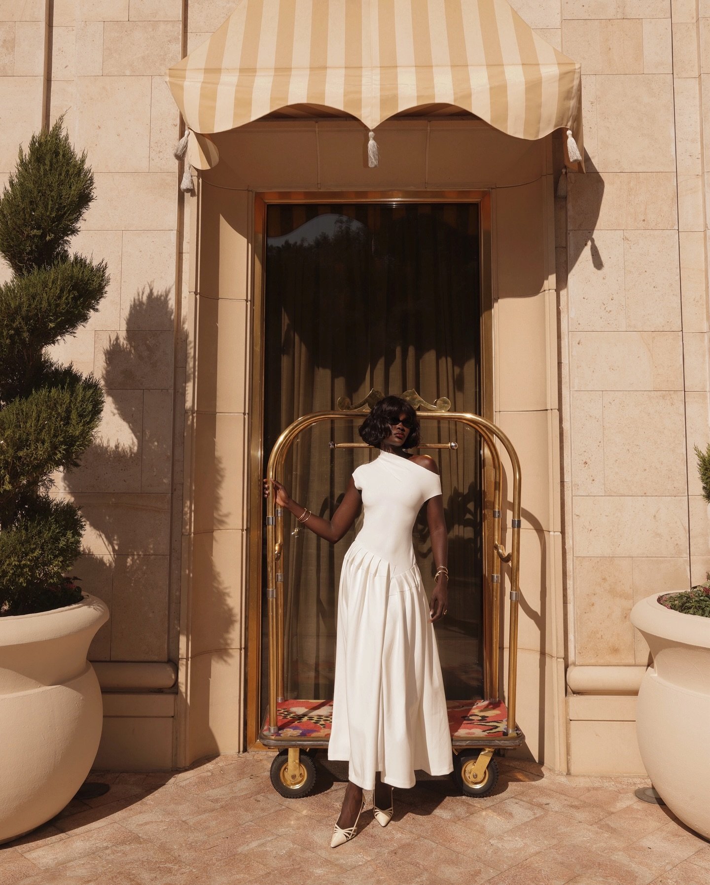 Modelo posa com vestido branco assimétrico de tecido fluido e sapato clássico e sofisticado em couro bege, em cenário