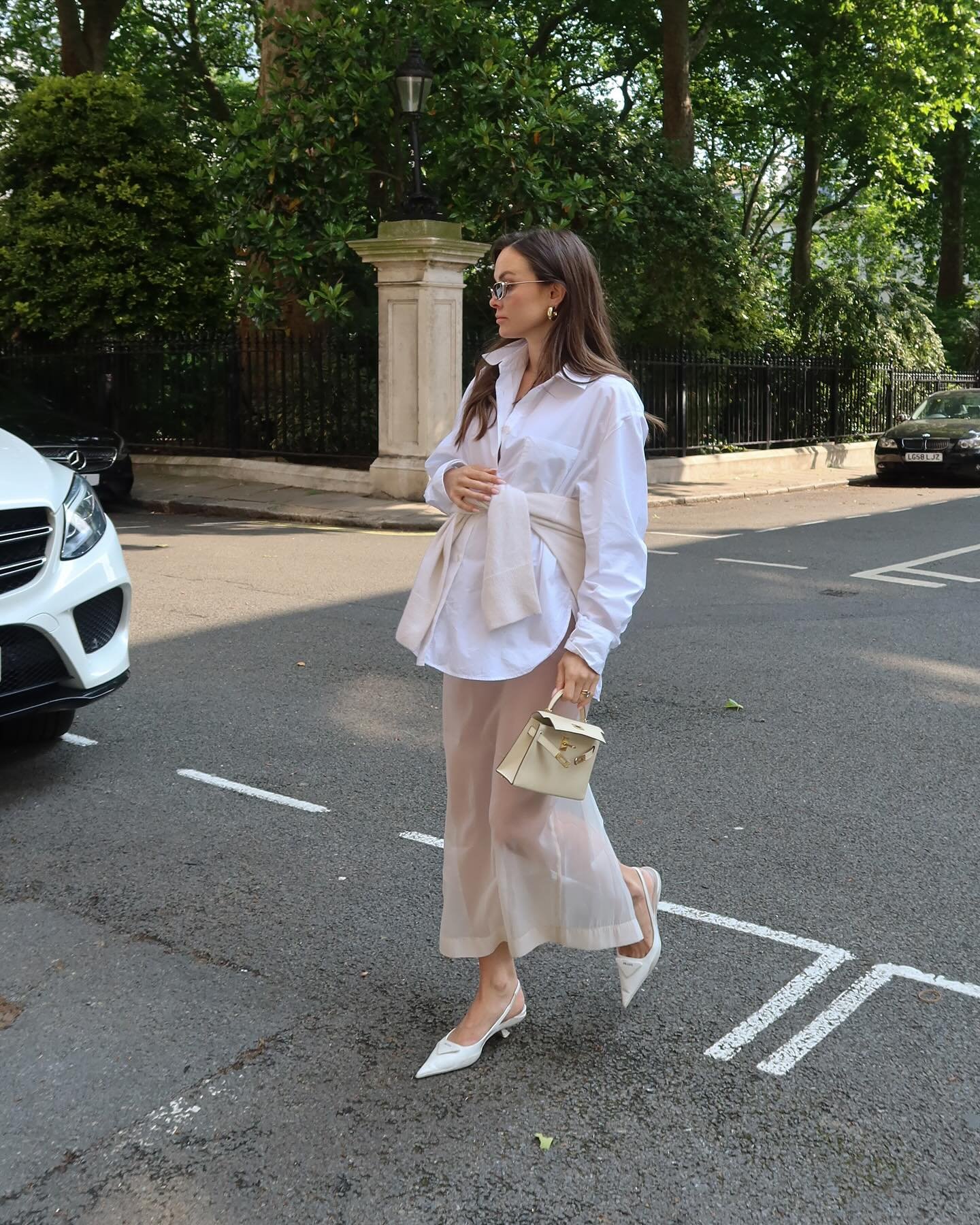Modelo caminha na rua com camisa branca oversized, saia midi nude fluida, bolsa bege estruturada e sapato clássico e