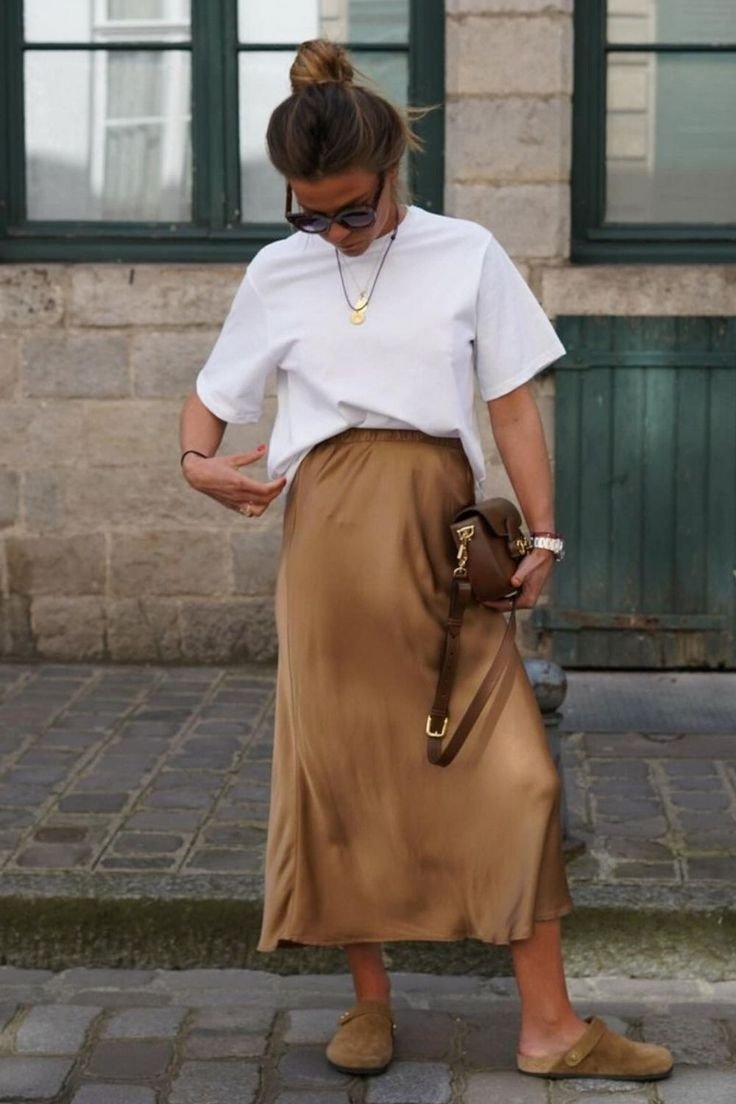 Modelo posa em look casual com clog em camurça caramelo, saia midi cetim dourada e camiseta branca oversized