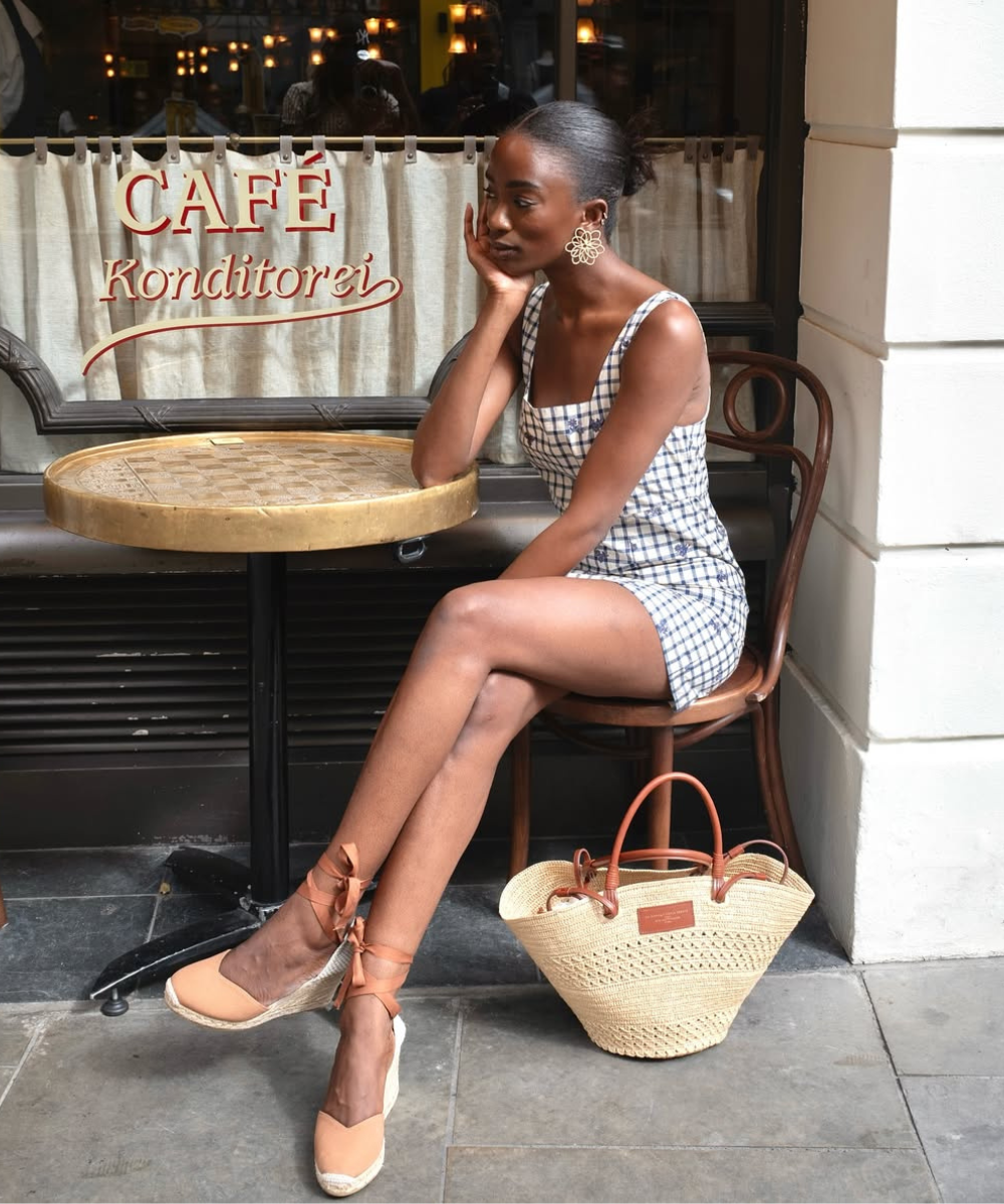 Modelo senta com postura pensativa usando vestido xadrez azul e branco, sapatos tendência para o verão 2026.