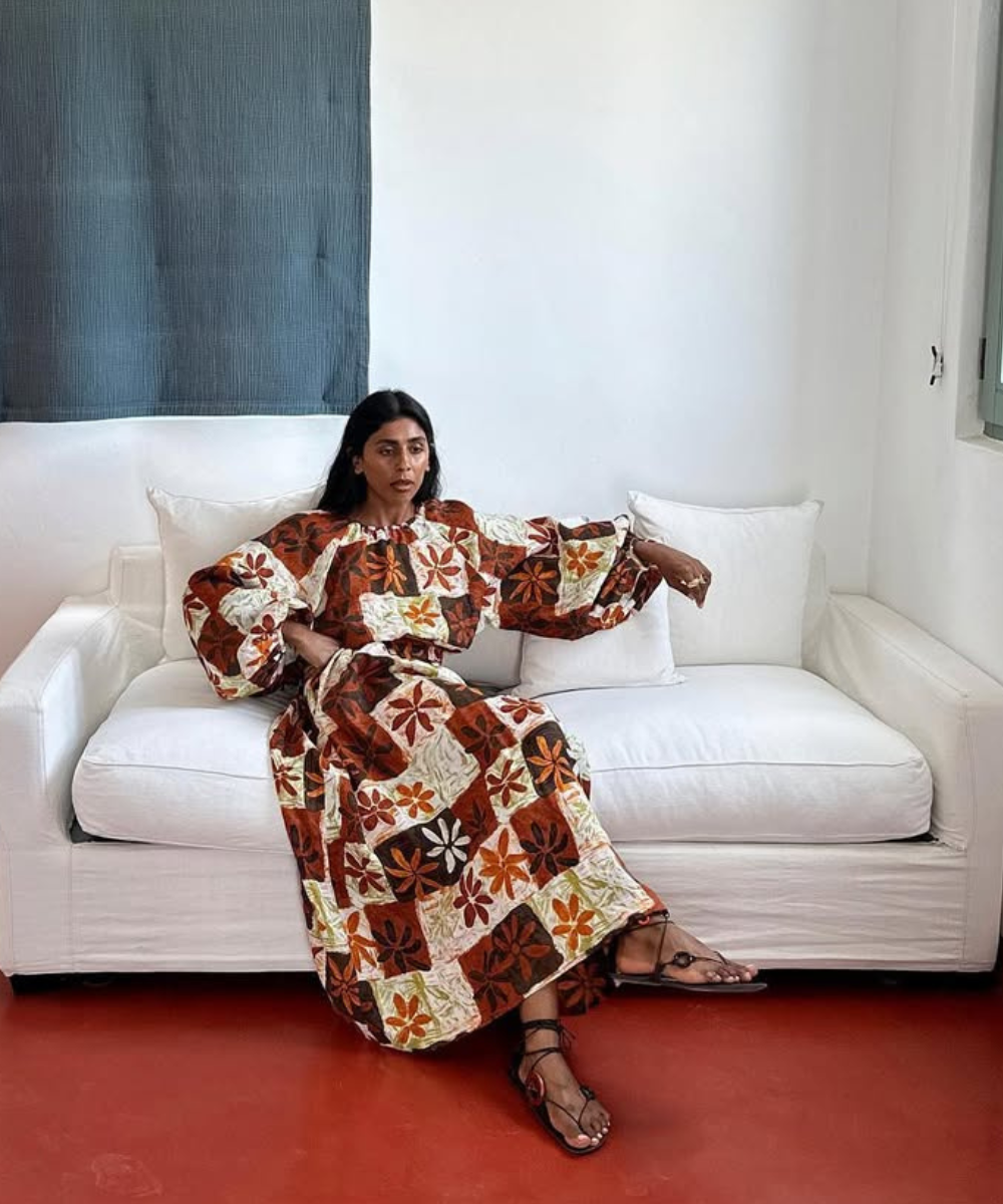 Modelo senta relaxada em sofá branco usando vestido longo estampado em tons terrosos e sapatos tendência para o verão 2026.