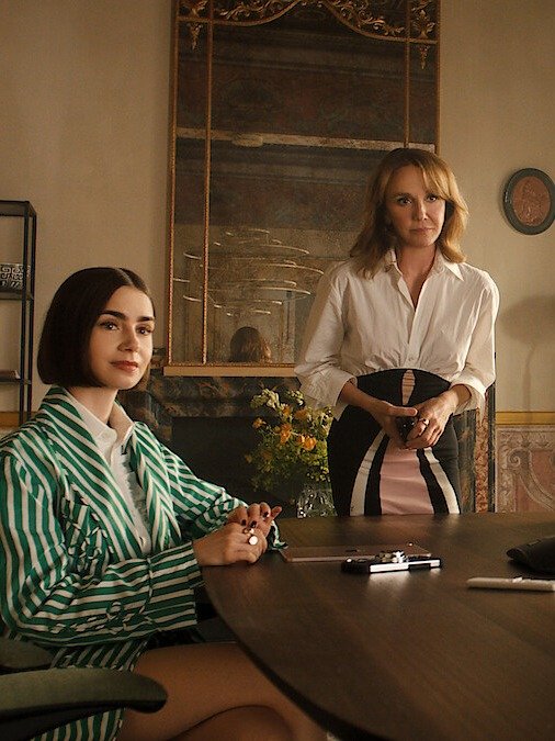 Duas mulheres em ambiente clássico; uma senta com blazer listrado verde e branco, a outra em pé com camisa branca e saia