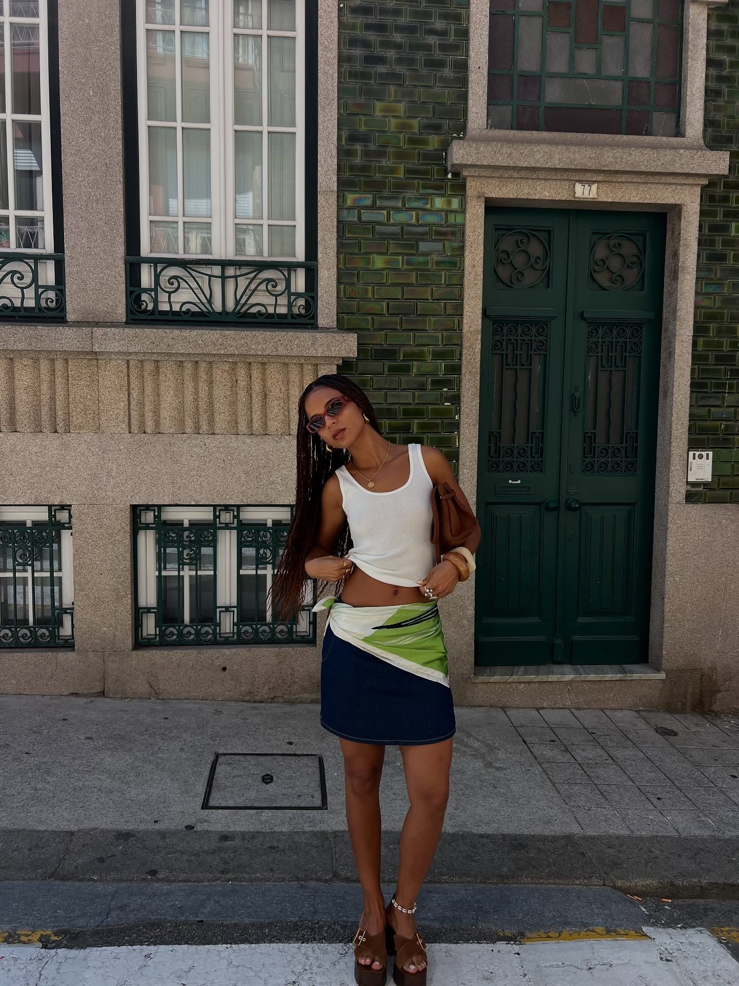 Modelo posa com tamanco marrom de salto bloco em look casual com regata branca, saia jeans e lenço verde, em cenário urbano