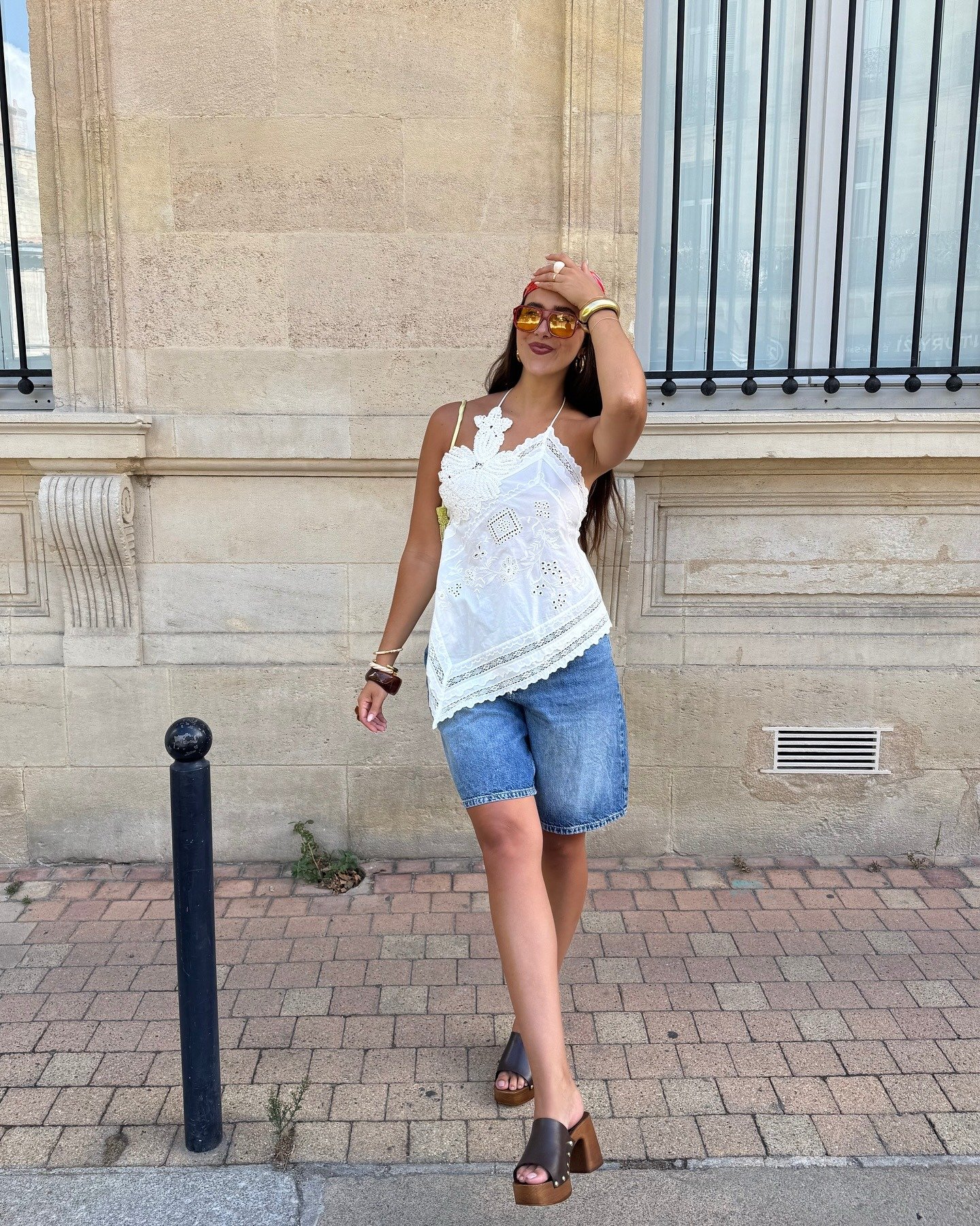 Modelo posa com tamanco marrom de salto bloco em madeira, bermuda jeans e blusa branca rendada, em clima descontraído.