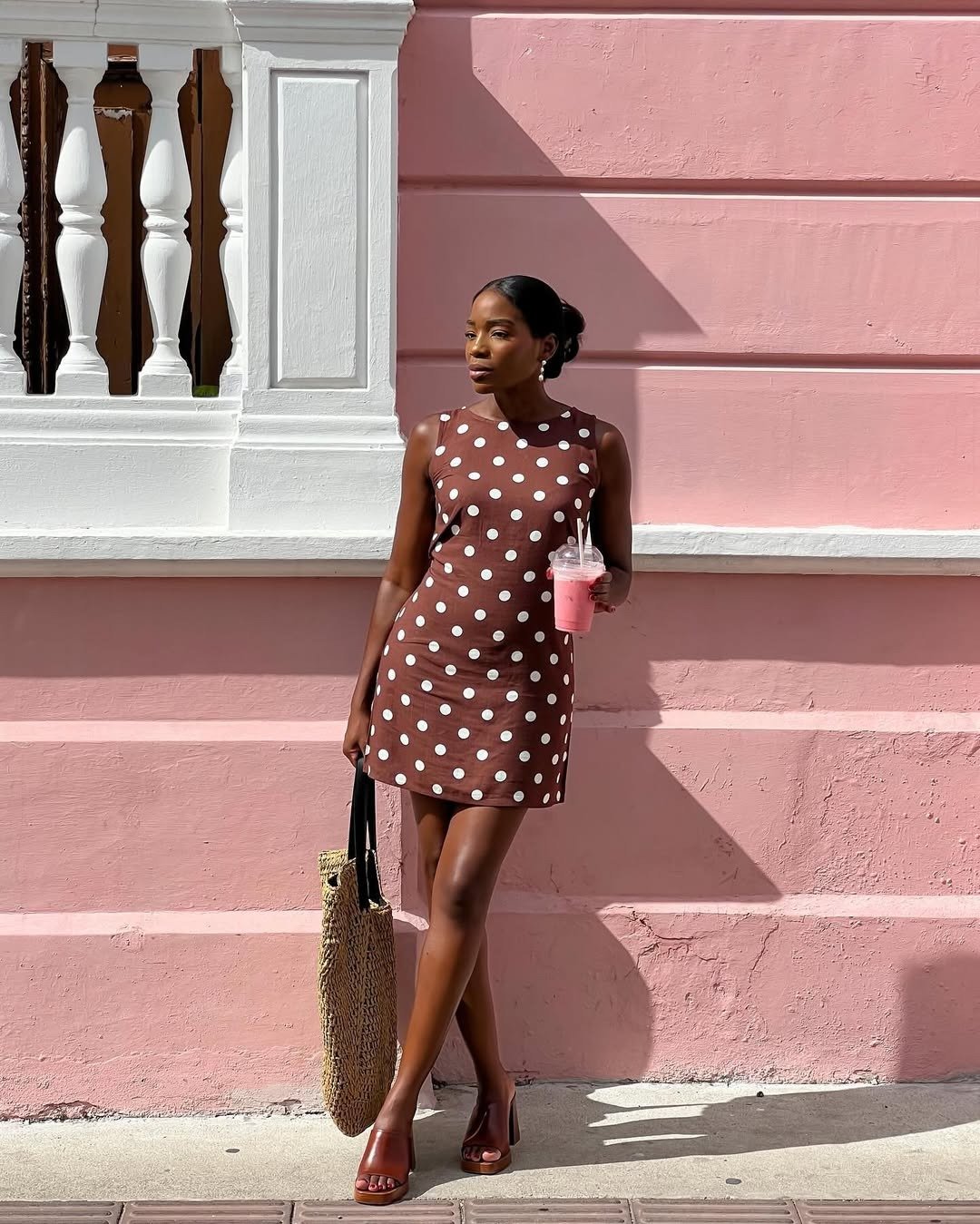 Modelo posa confiante com vestido marrom de bolinhas brancas, tamanco marrom em couro e bolsa de palha, em cenário rosa