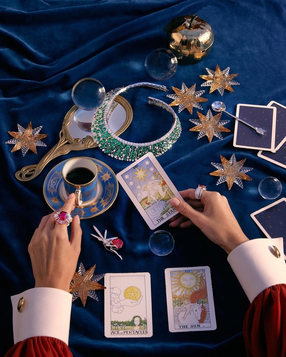 Mãos com anéis de pedras vermelhas seguram carta do tarot de 2026 sobre veludo azul, cercadas por joias e estrelas douradas.