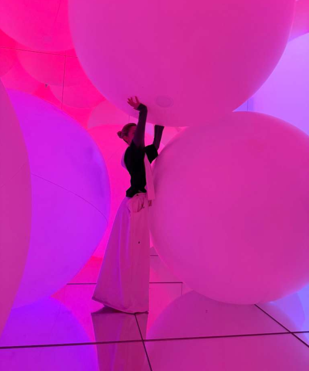Modelo em Tóquio interage com grandes esferas iluminadas em tons vibrantes de rosa e roxo, usando blusa preta justa e calça
