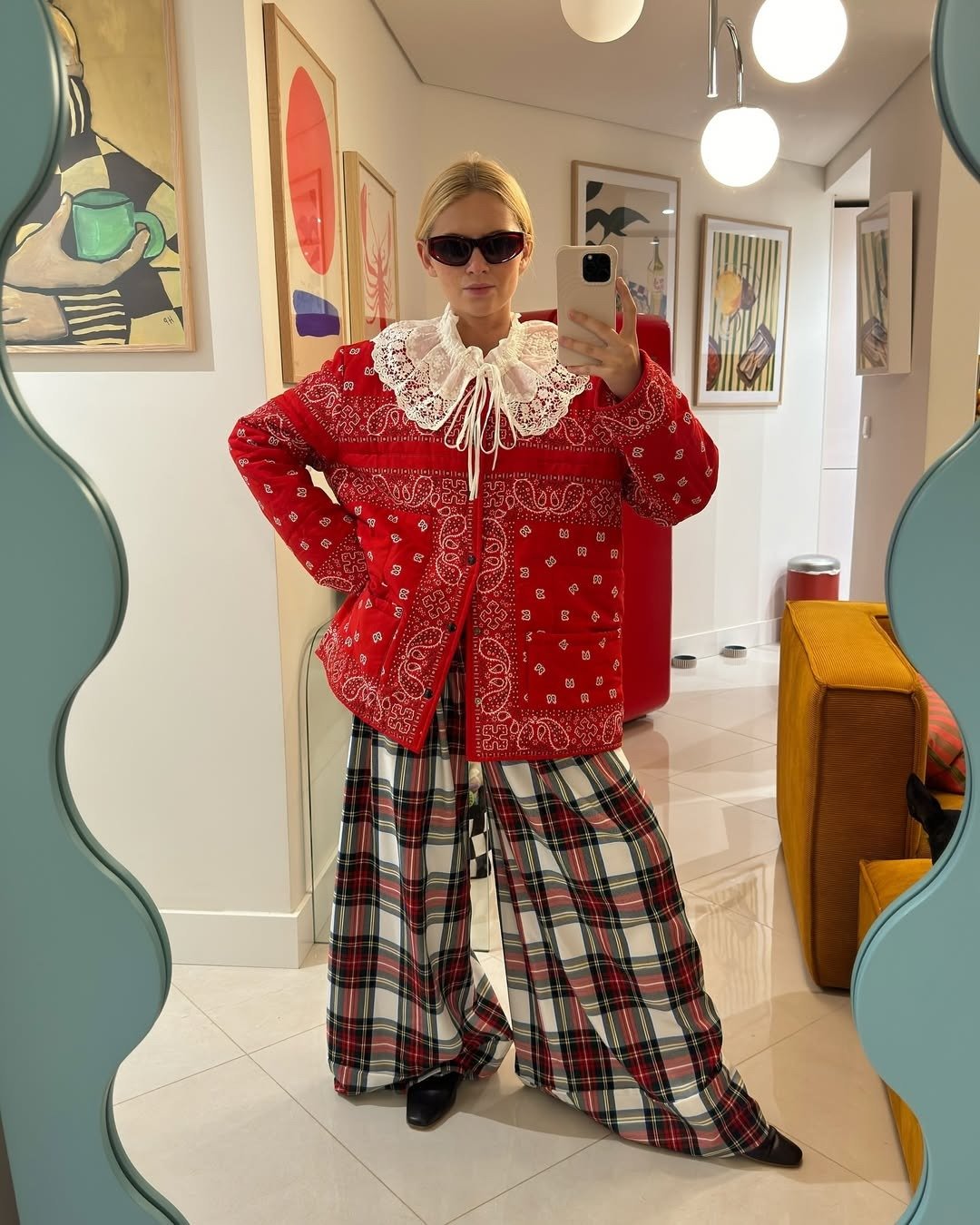 Modelo posa com jaqueta vermelha estampada, gola de renda branca e calça xadrez ampla, expressando as tendências de moda de