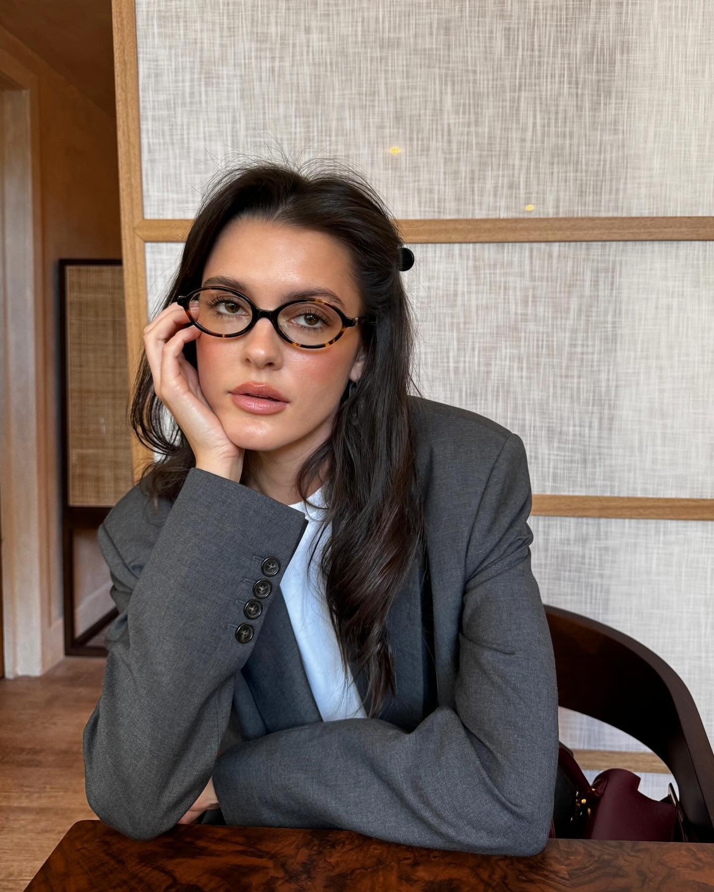 Modelo posa com blazer cinza oversized e camiseta branca, óculos de armação tartaruga e expressão serena, refletindo