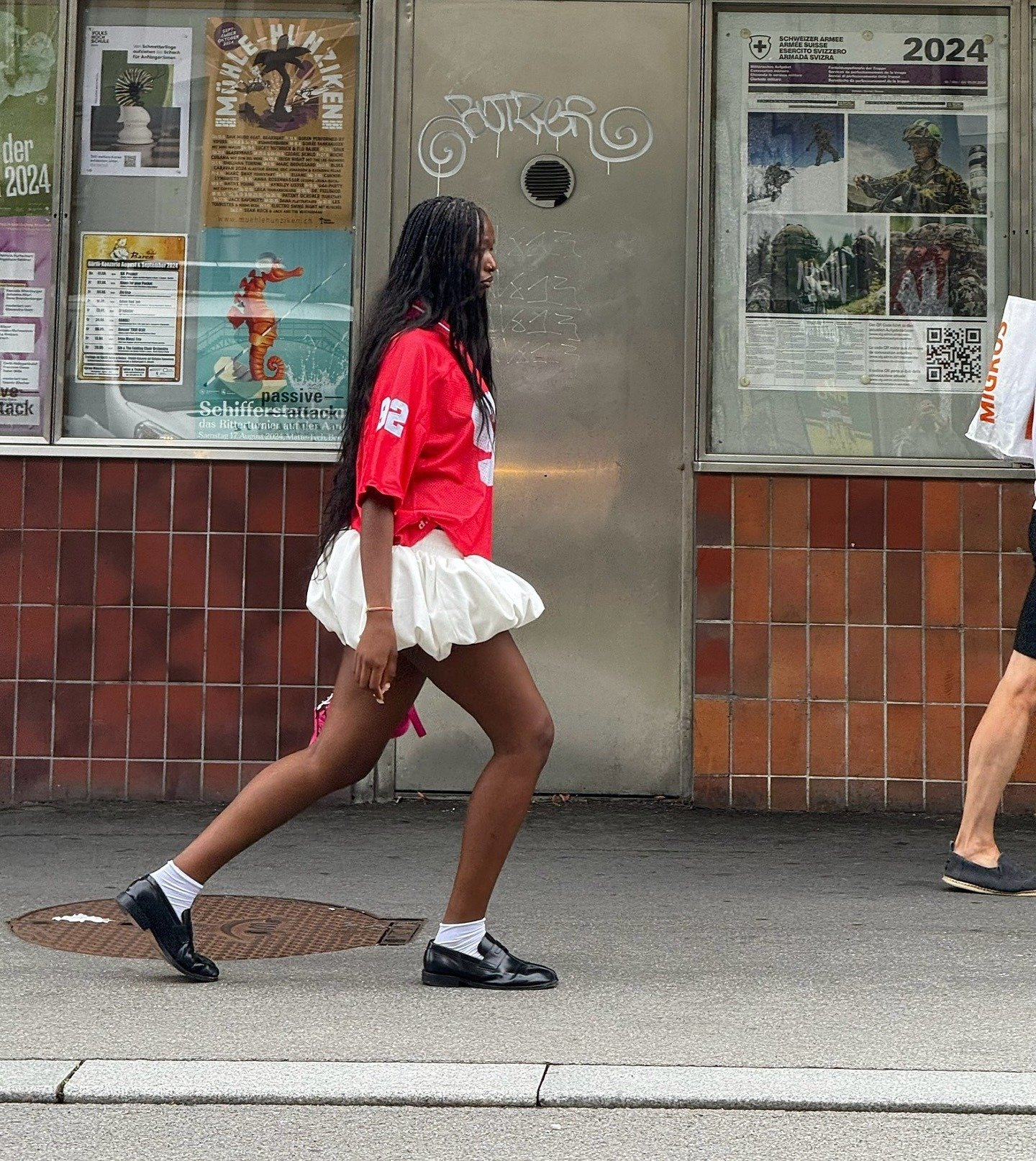 Modelo caminha com postura decidida usando camiseta oversized vermelha, saia bufante branca e sapatos pretos, refletindo