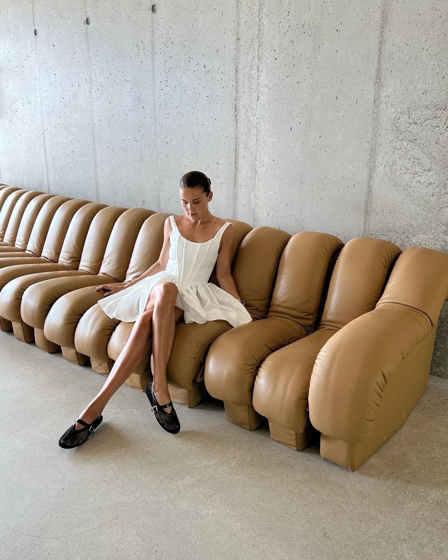 Modelo senta com postura relaxada em sofá caramelo, usando vestido branco estruturado e sapatilhas pretas, refletindo