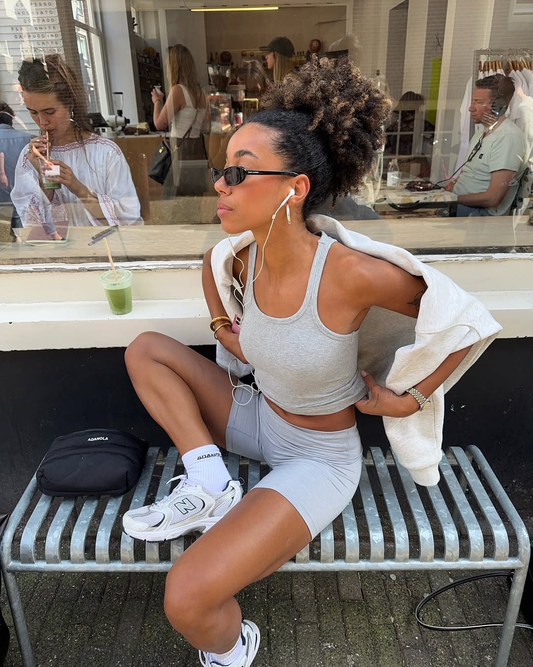 Mulher senta em banco metálico com tênis esportivo branco New Balance, shorts e regata cinza, óculos escuros e cabelo preso