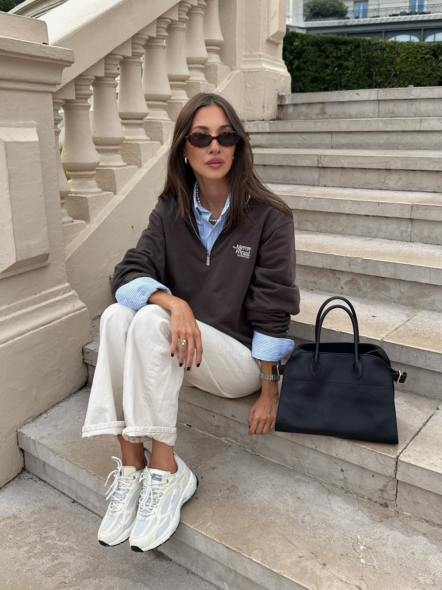 Mulher sentada em escada usa tênis esportivo branco com detalhes em cinza, calça clara ampla, camisa listrada e moletom