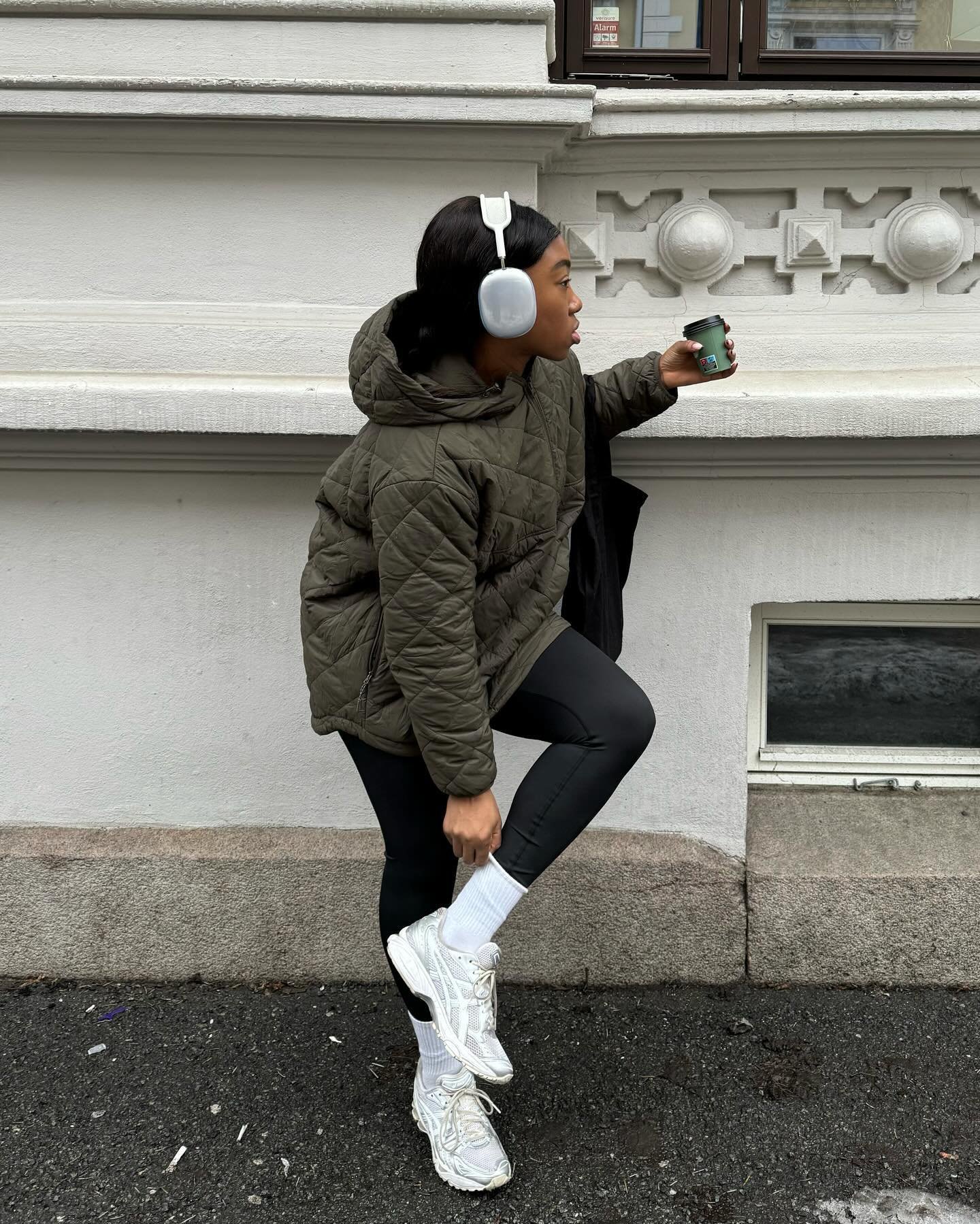 Modelo ajusta o tênis esportivo branco de solado robusto, combinando com legging preta e jaqueta matelassê verde-oliva em