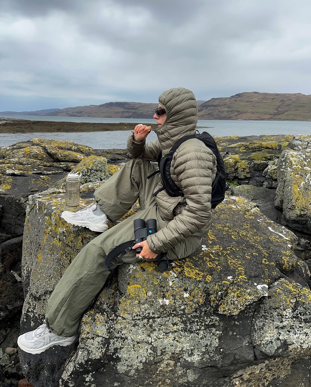 Modelo senta sobre pedras cobertas de líquens com postura contemplativa, vestindo tênis esportivo branco robusto, calça