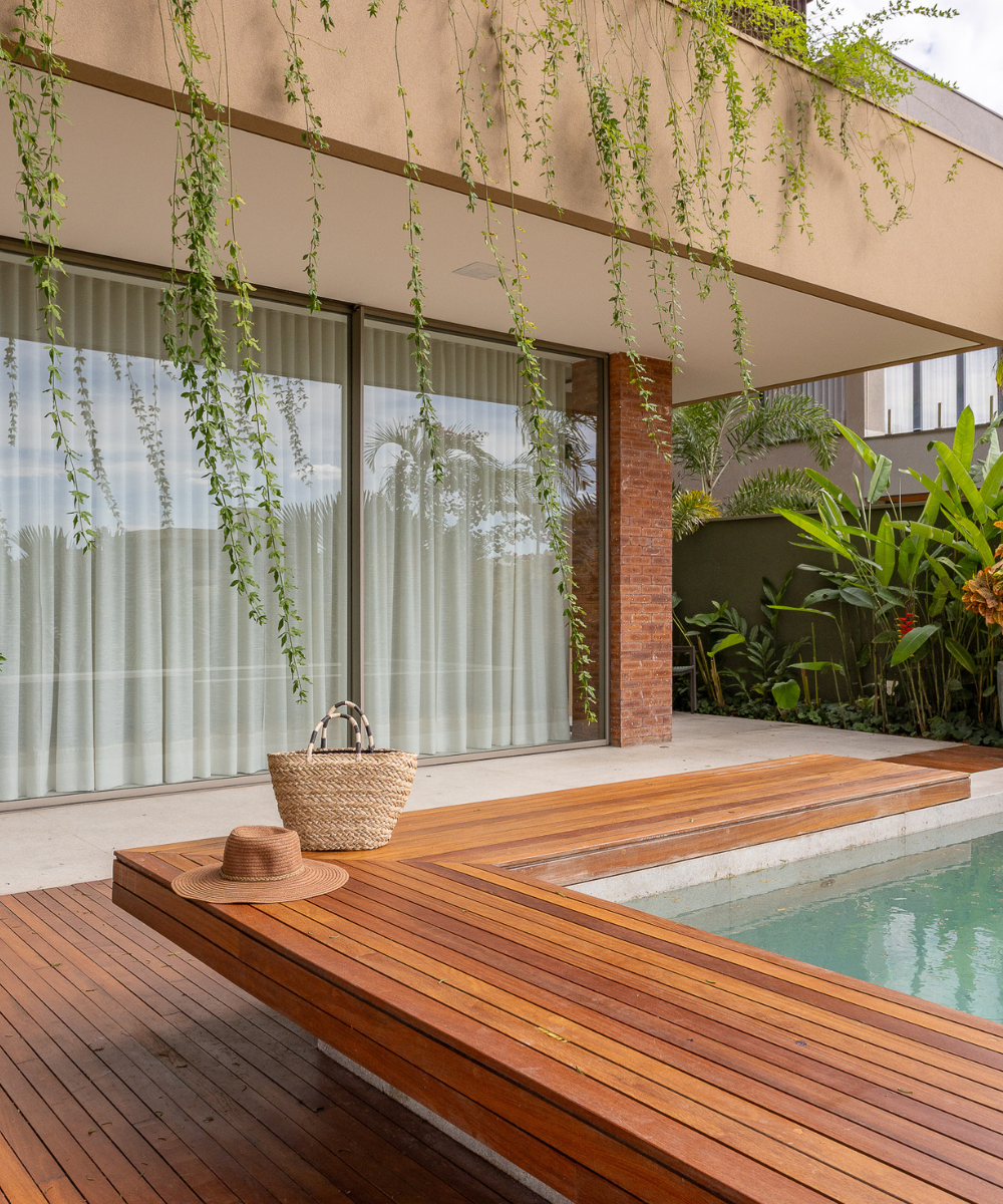 Varanda com piscina exibe parede de tijolinho aparente em tom terroso, deck de madeira e vegetação suspensa, criando