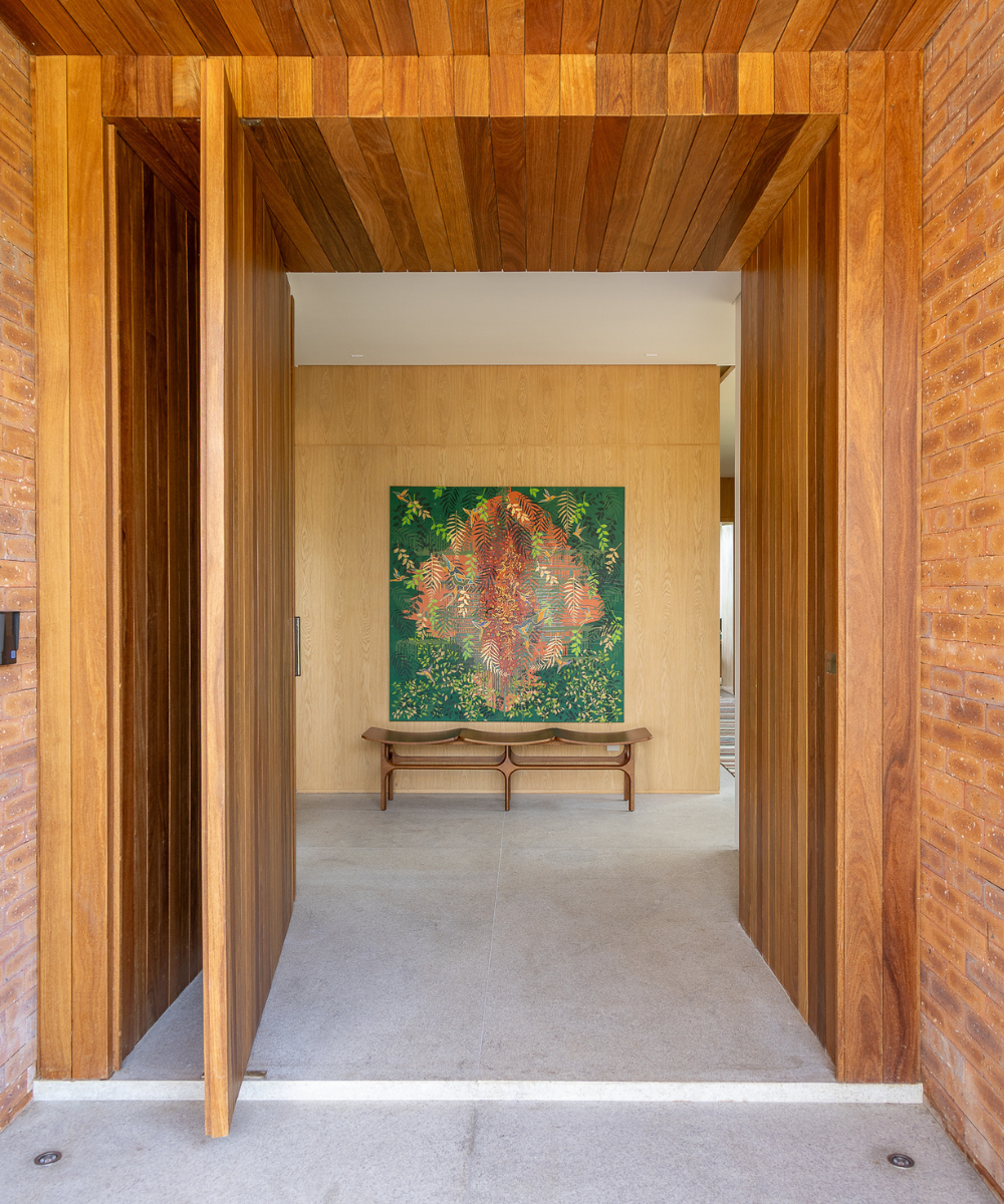 Entrada com parede de tijolinho aparente em tons terrosos, porta pivotante e teto em madeira, criando ambiente acolhedor e