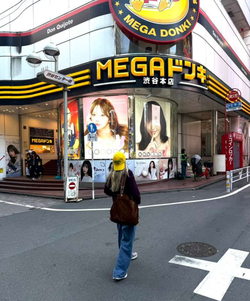 Mulher caminha em Tóquio usando boné amarelo, casaco escuro e calça jeans ampla, diante da fachada vibrante da Mega Donki,