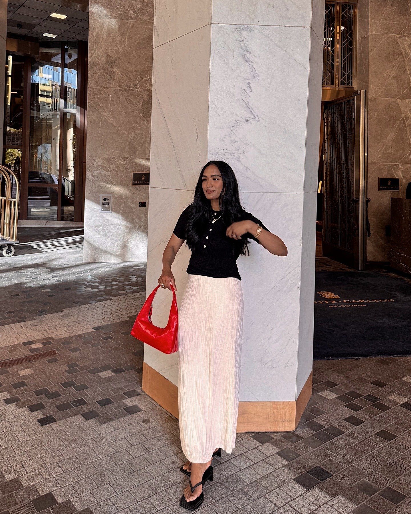 Modelo posa com blusa preta de botões, saia longa plissada off-white e bolsa vermelha, evidenciando tendências de verão