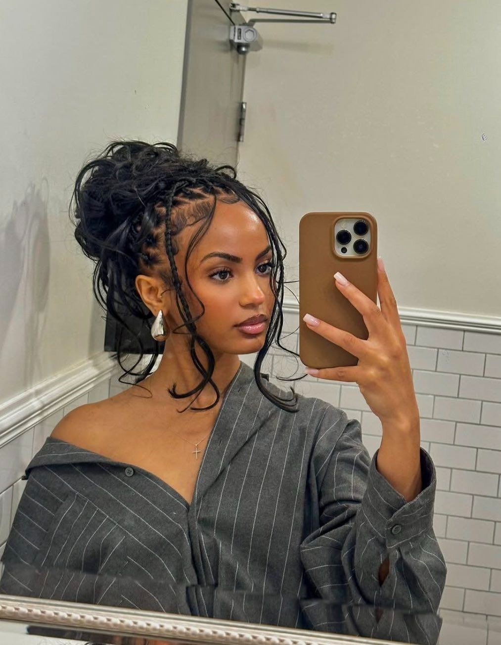 Modelo posa para selfie com penteados com trança presos em coque bagunçado, blusa cinza listrada e acessórios delicados.