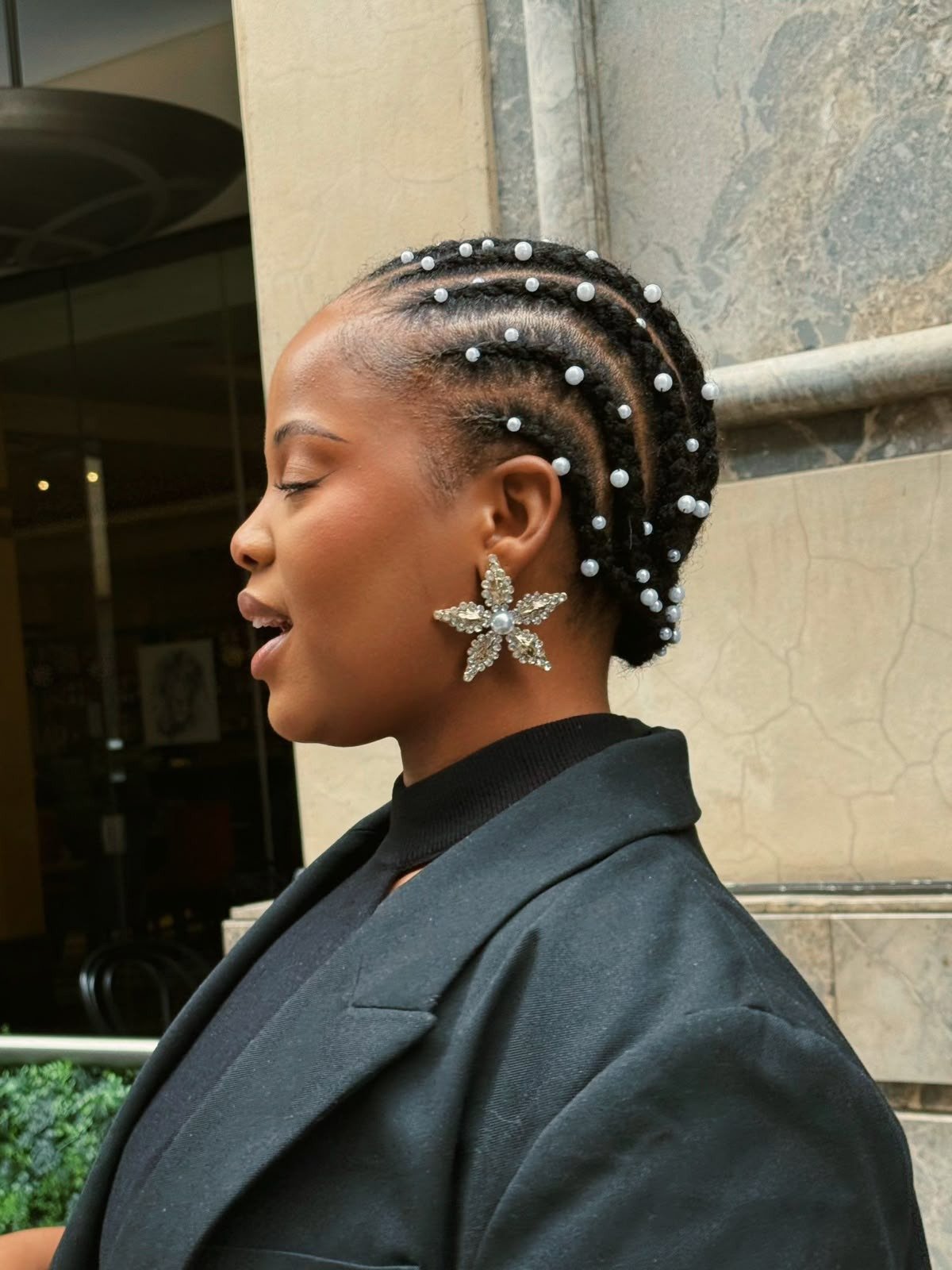 Modelo posa de perfil com penteados com trança adornados por pérolas, usando blazer preto e brinco floral brilhante.
