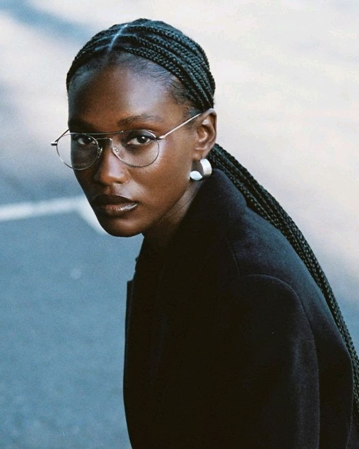 Modelo com penteados com trança em microtranças presas, usa blazer preto, óculos de armação fina e brincos prateados.