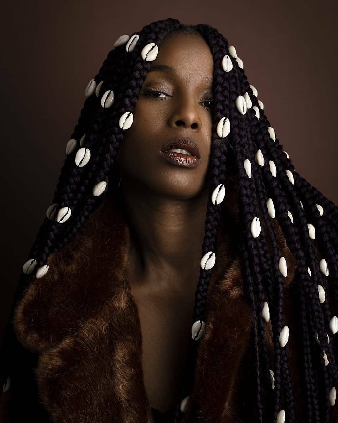 Modelo posa com penteados com trança longas e volumosas adornadas por búzios brancos, usando casaco marrom.