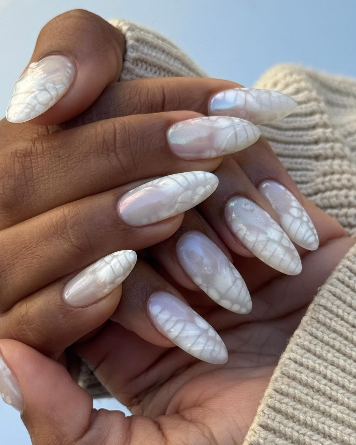 Unhas brancas alongadas em formato amendoado com efeito perolado e texturas delicadas, mãos envoltas em malha bege.