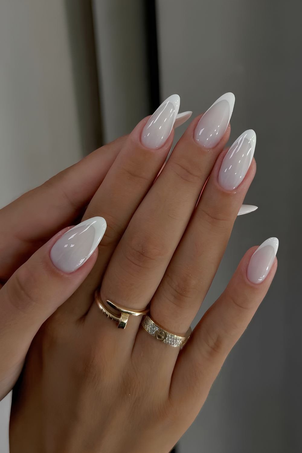 Mãos femininas exibem unhas brancas em formato amendoado com acabamento glossy, acompanhadas por anéis dourados delicados.