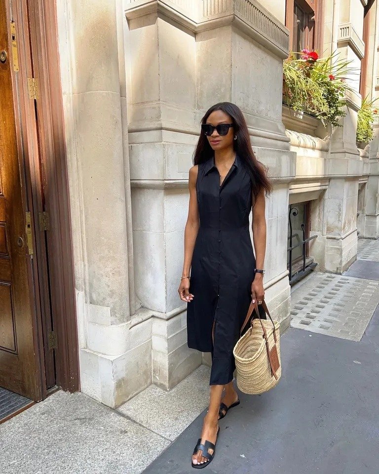 Mulher caminha com vestido preto midi sem mangas e rasteirinha preta, combinando bolsa de palha em cenário urbano elegante.