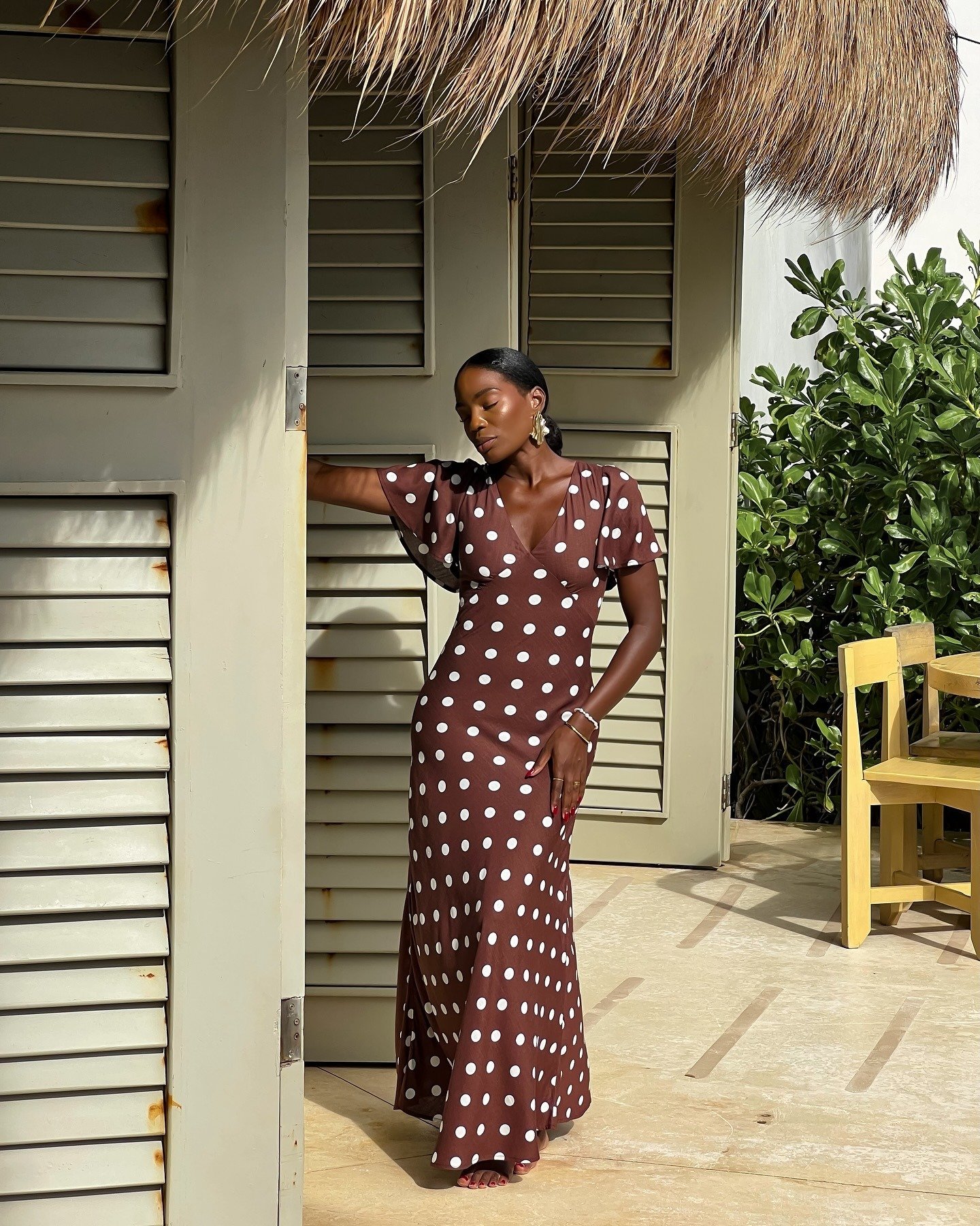 Modelo posa com vestido longo marrom de poás brancos, decote em V e mangas curtas fluídas, combinando com rasteirinha nude