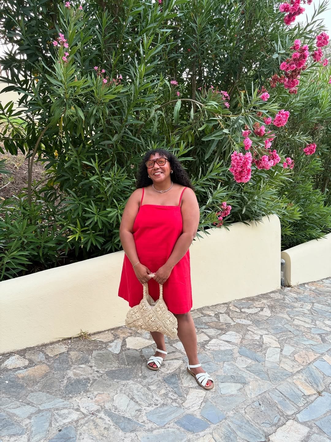 Modelo sorri e posa com vestido vermelho de alças finas em tecido leve, combinando rasteirinha branca e bolsa de crochê