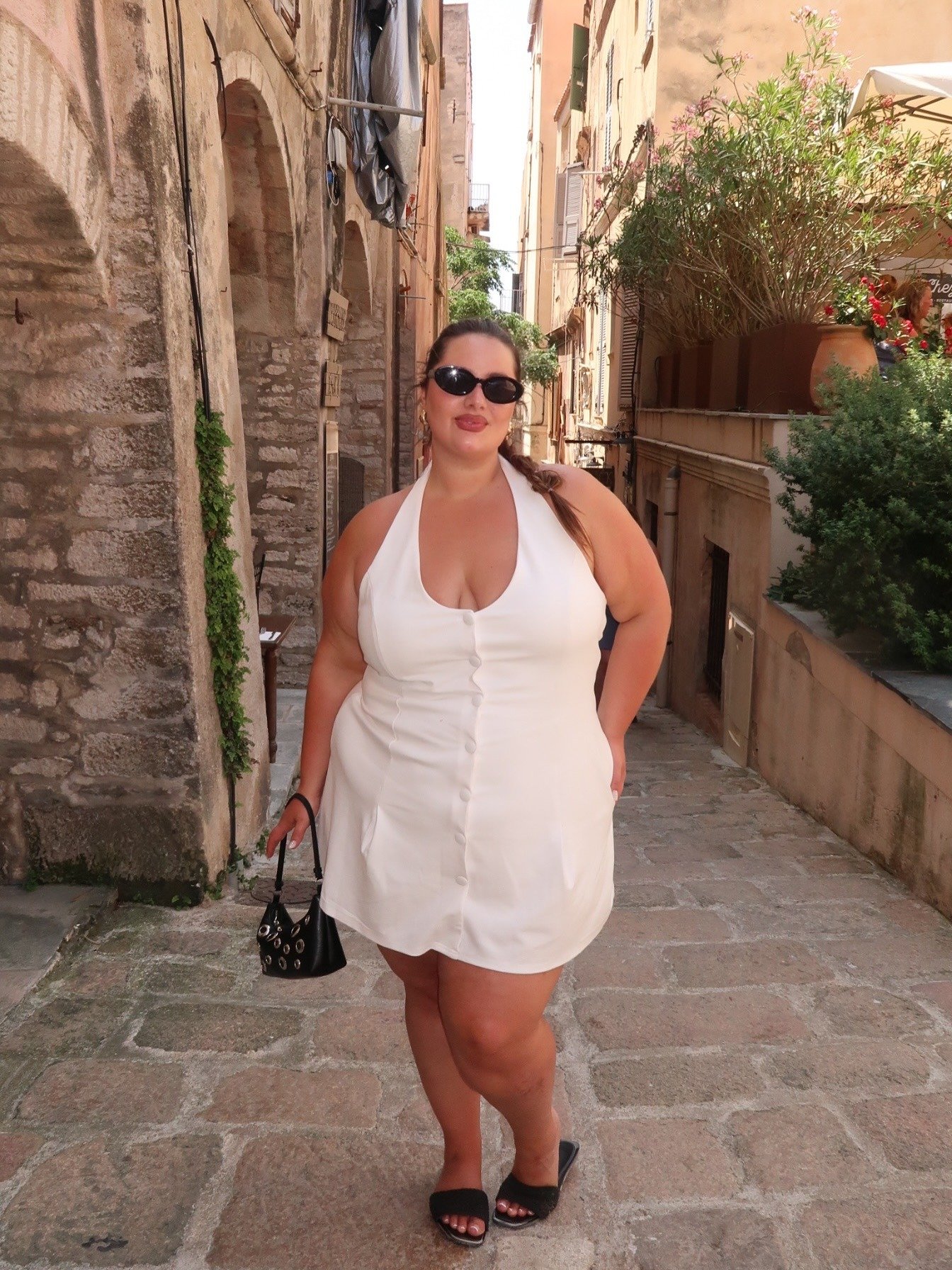 Modelo posa com vestido frente única branco de tecido liso, botões frontais e saia curta, combinando com bolsa preta.