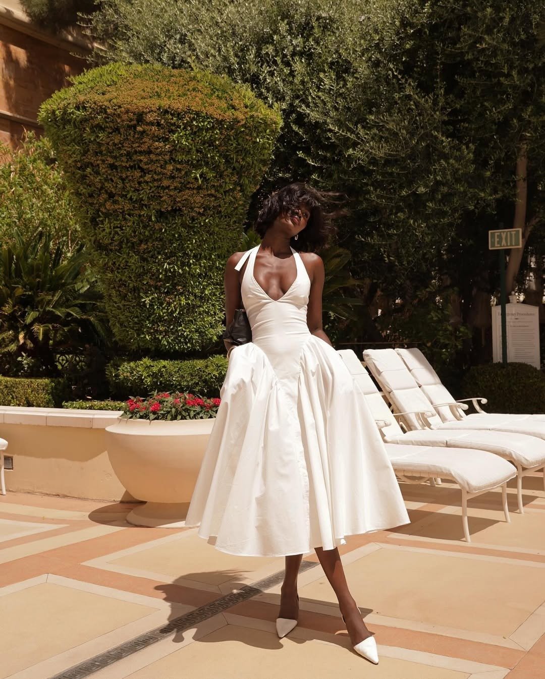 Modelo posa com vestido frente única branco de tecido estruturado, saia rodada e decote profundo, em ambiente ensolarado.