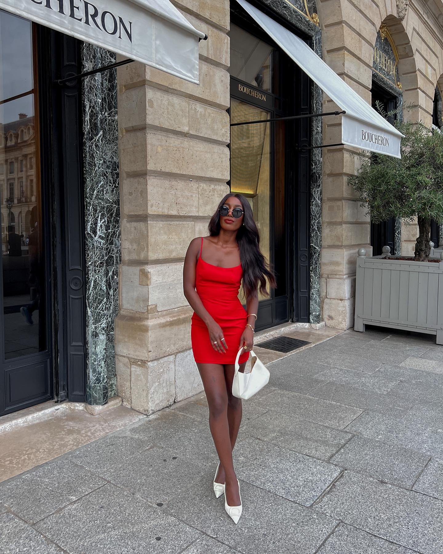 Modelo posa com vestido vermelho justo de alças finas, combinando bolsa e scarpin branco, em cenário urbano elegante.