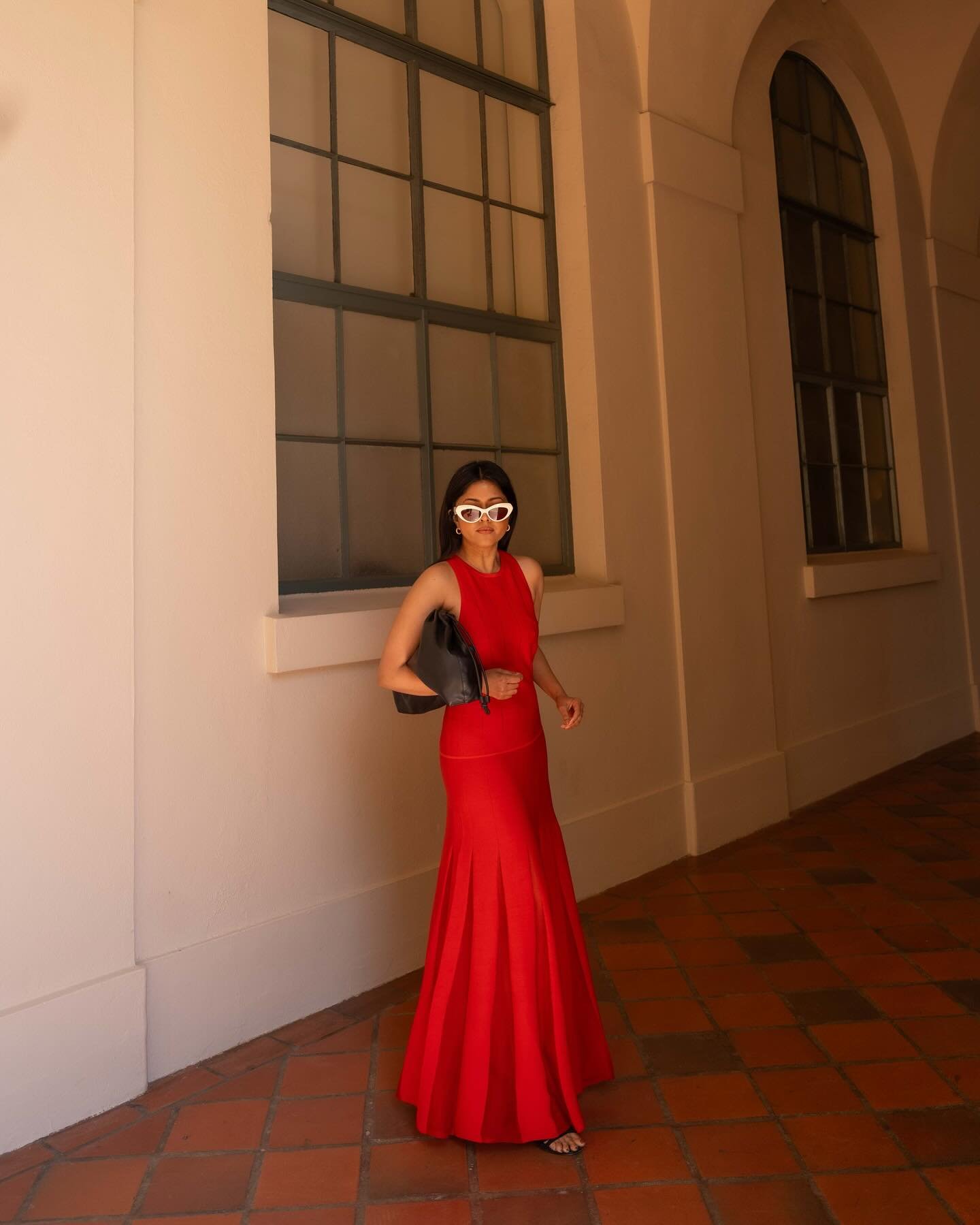 Modelo caminha com vestido vermelho longo justo ao corpo e saia flare, combinando bolsa preta e óculos branco em ambiente