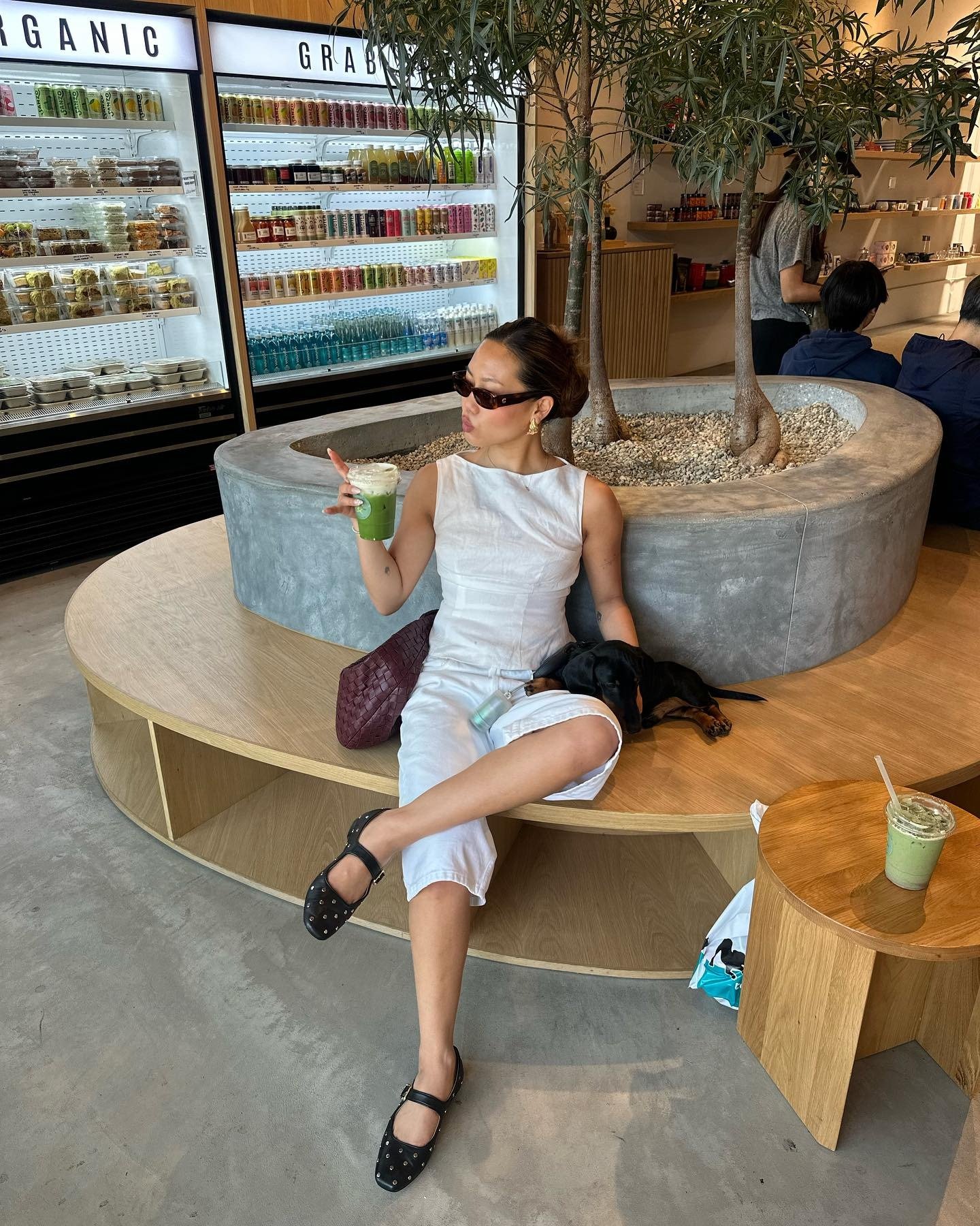 Modelo posa sentada com vestido branco de linho, sapatos pretos com tachas e acessórios minimalistas