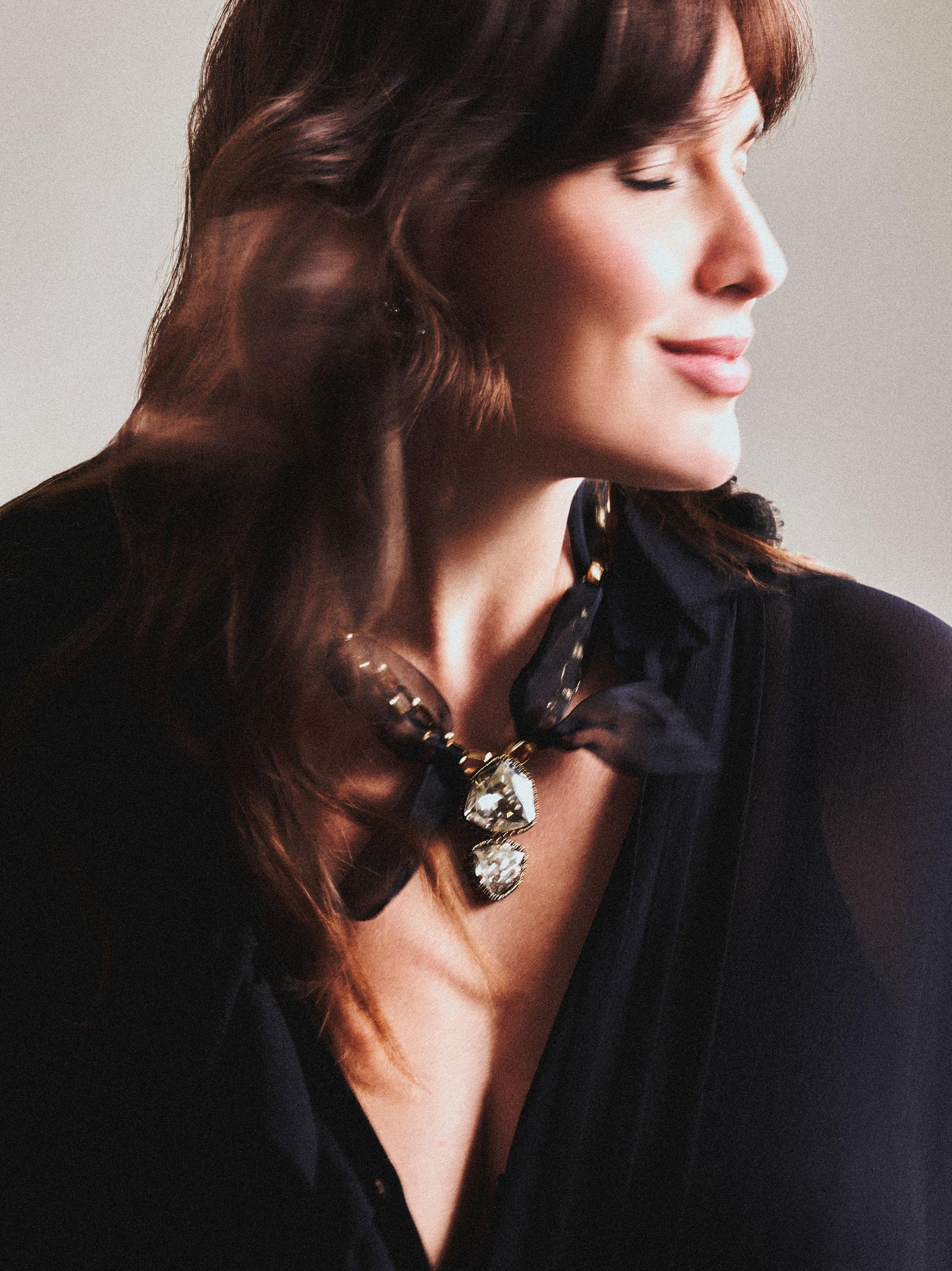 Bia Perotti posa de perfil com expressão serena, usando blusa preta de tecido leve e colar statement com pedras facetadas e