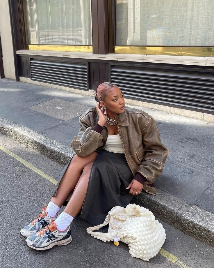 Looks com tênis esportivo New Balance branco e azul, saia midi preta, jaqueta oversized marrom e acessórios prateados, modelo