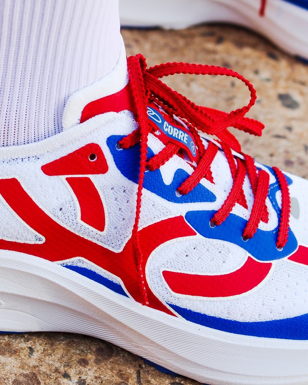 Tênis Corre 4 em tecido mesh branco com detalhes em vermelho e azul, cadarços vermelhos e design esportivo vibrante.