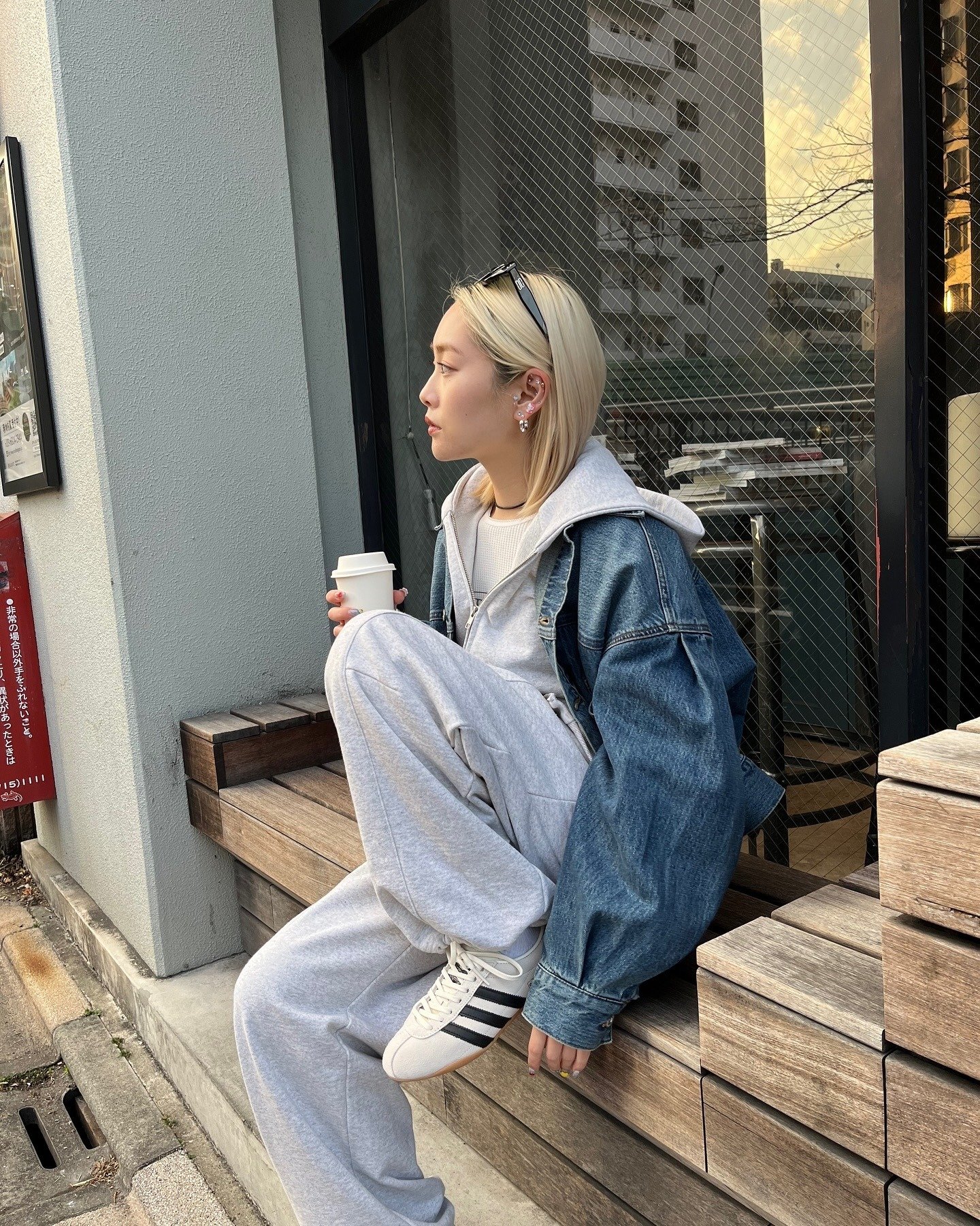 Jovem sentada em banco de madeira com moletom cinza, jaqueta jeans oversized e tênis adidas tokyo branco com listras pretas,