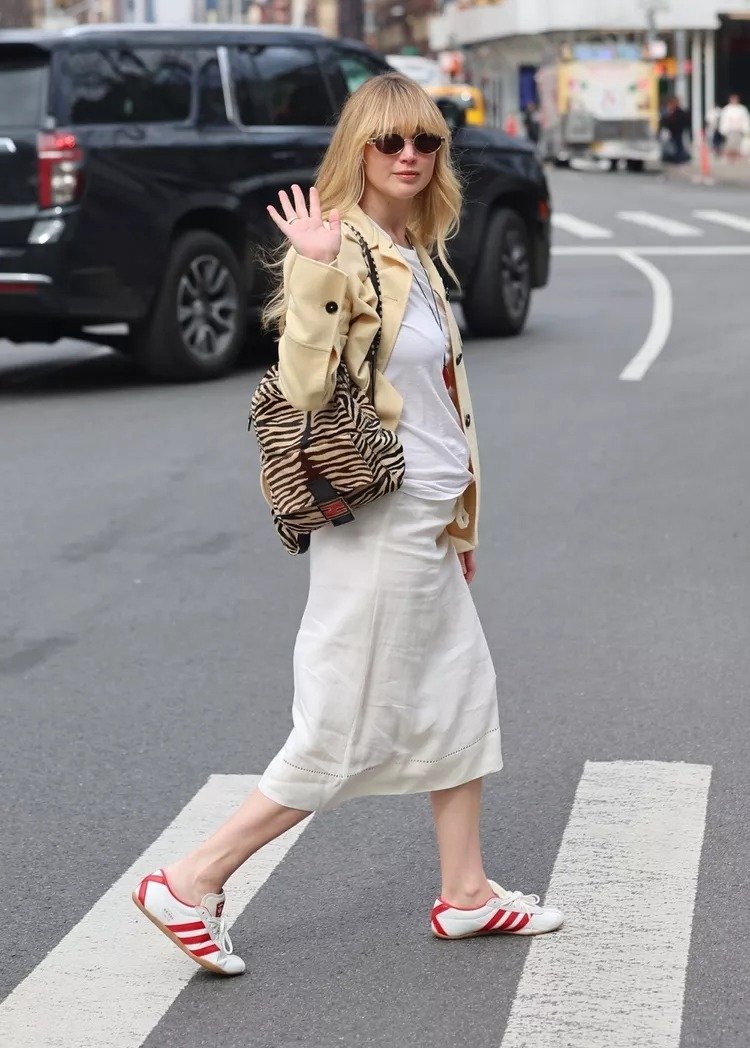 Modelo caminha na rua com tênis adidas tokyo branco e vermelho, saia midi branca, camiseta básica e jaqueta bege, estilo