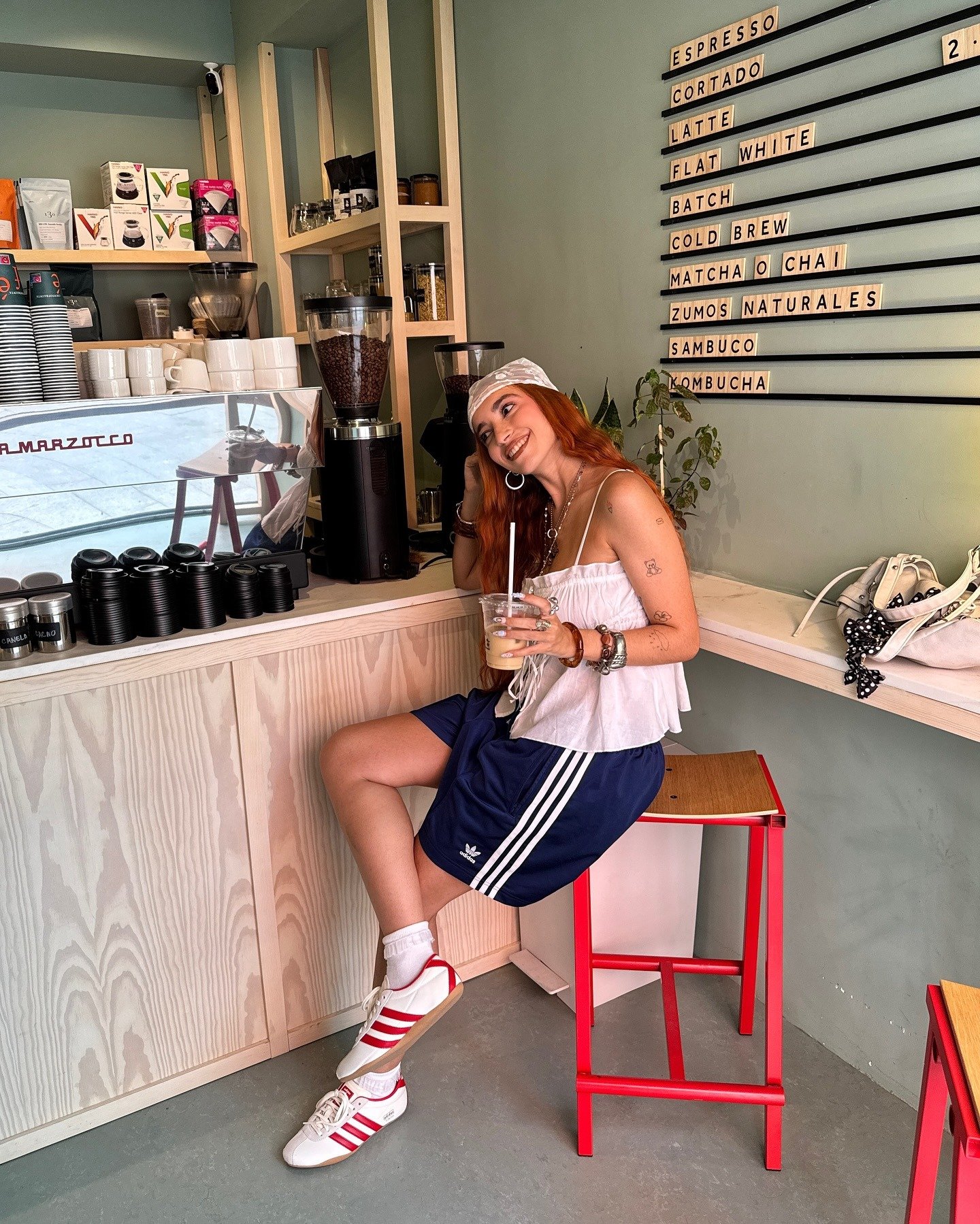 Modelo sentada em banco vermelho em café, veste shorts adidas Tokyo azul marinho com listras brancas, tênis branco e
