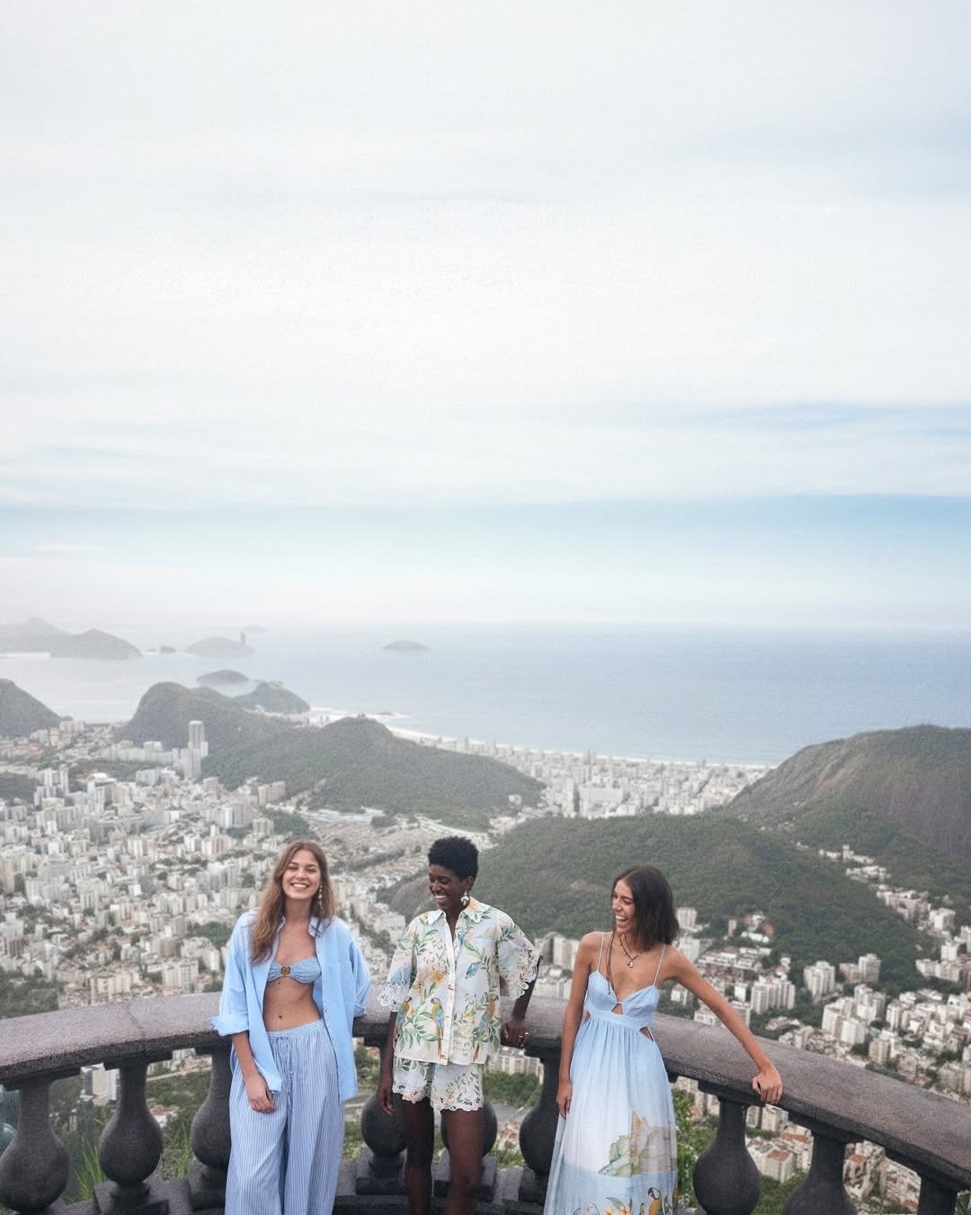 Três mulheres posam sorrindo com peças fluidas e leves em tons de azul e estampas naturais, expressando liberdade e conexão