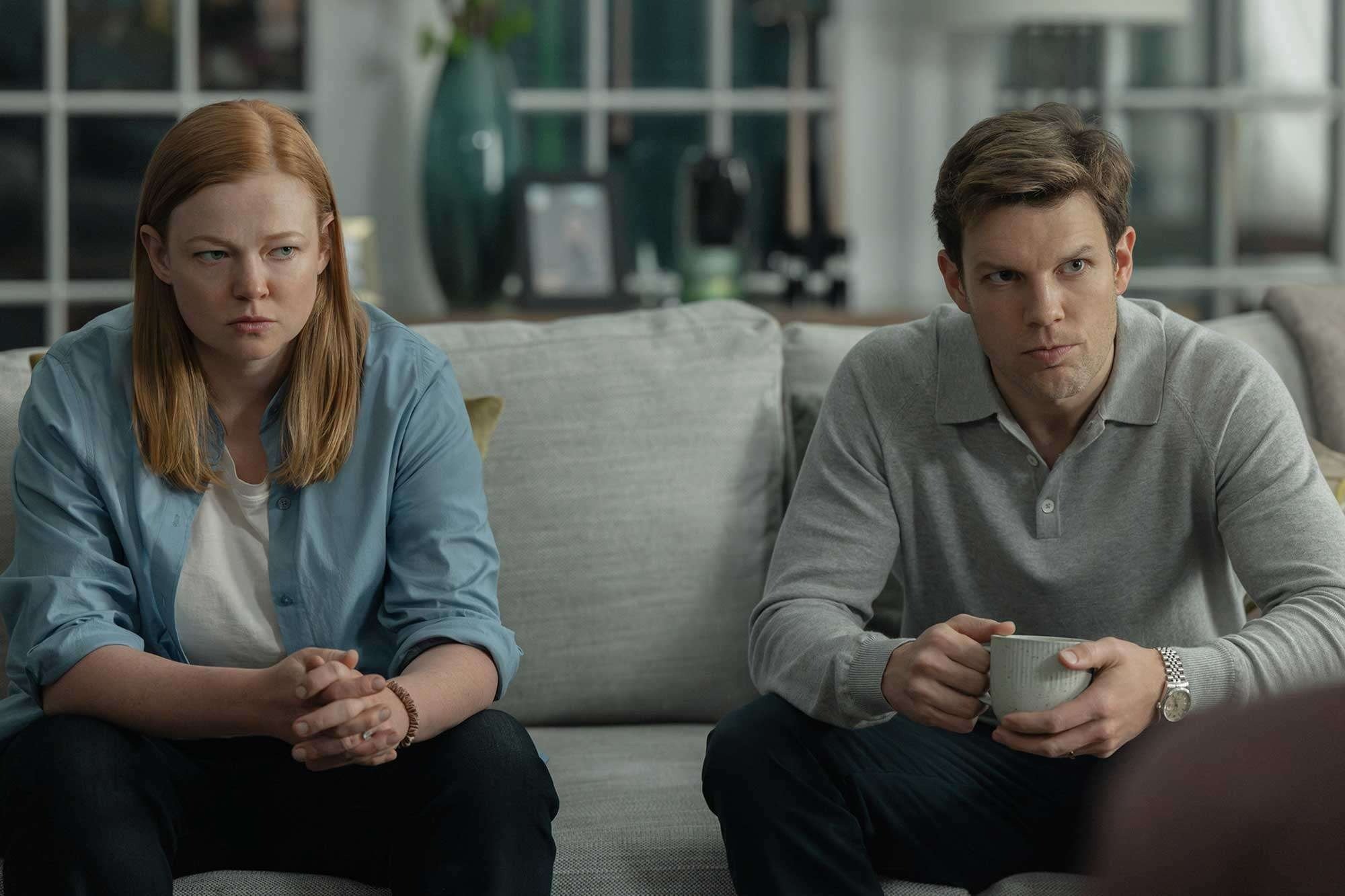 Cena de All Her Fault mostra mulher de camisa azul clara sobre camiseta branca e homem com suéter cinza, ambos sentados