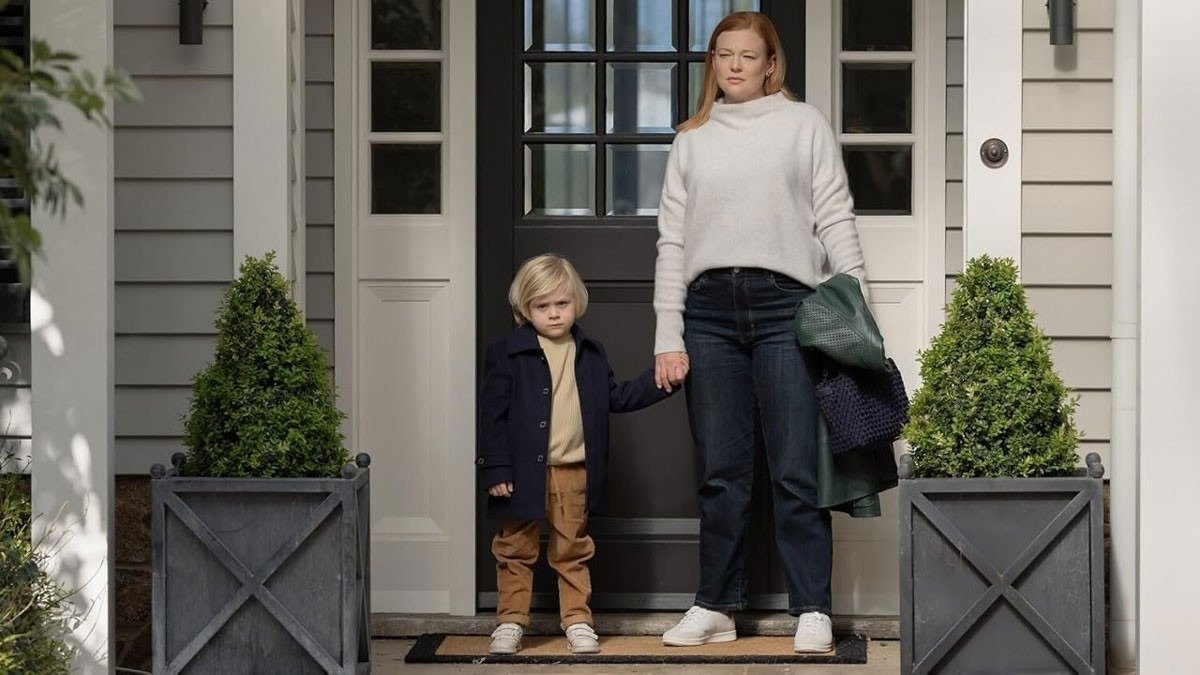 Mulher com suéter branco de gola alta e jeans escuros segura a mão do menino de casaco azul marinho. All Her Fault