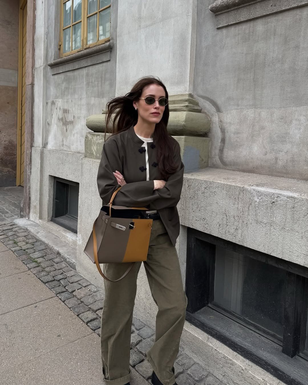 Modelo posa com jaqueta marrom de modelagem reta e detalhes em nó chinês, combinada a calça verde militar e bolsa bicolor,