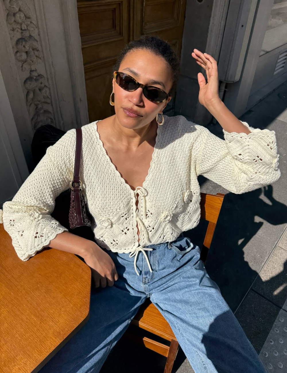 Modelo posa sentada ao sol usando bata feminina de tricô off-white com amarração frontal e mangas flare, combinada com jeans.