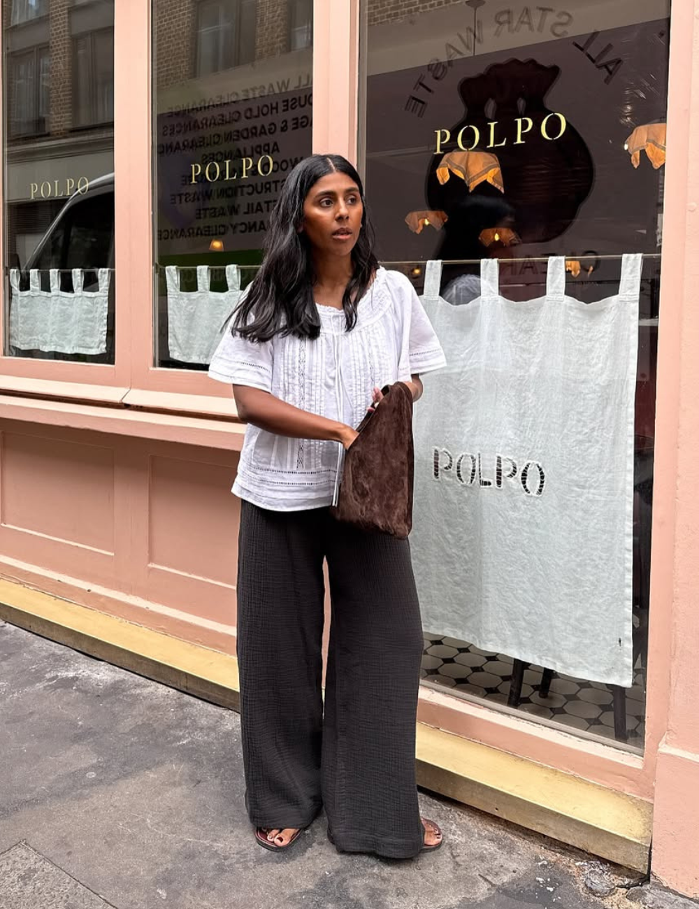 Mulher posa com bata feminina branca de algodão com bordados delicados, combinada a calça preta ampla e bolsa marrom.