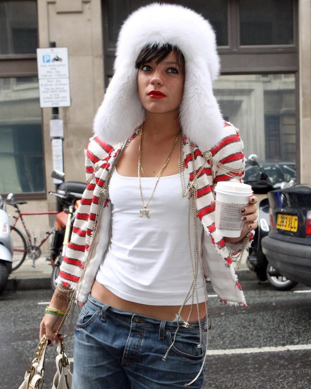 Lily Allen posa na rua com jaqueta listrada vermelha e branca em tecido texturizado, regata branca, jeans e chapéu de pele